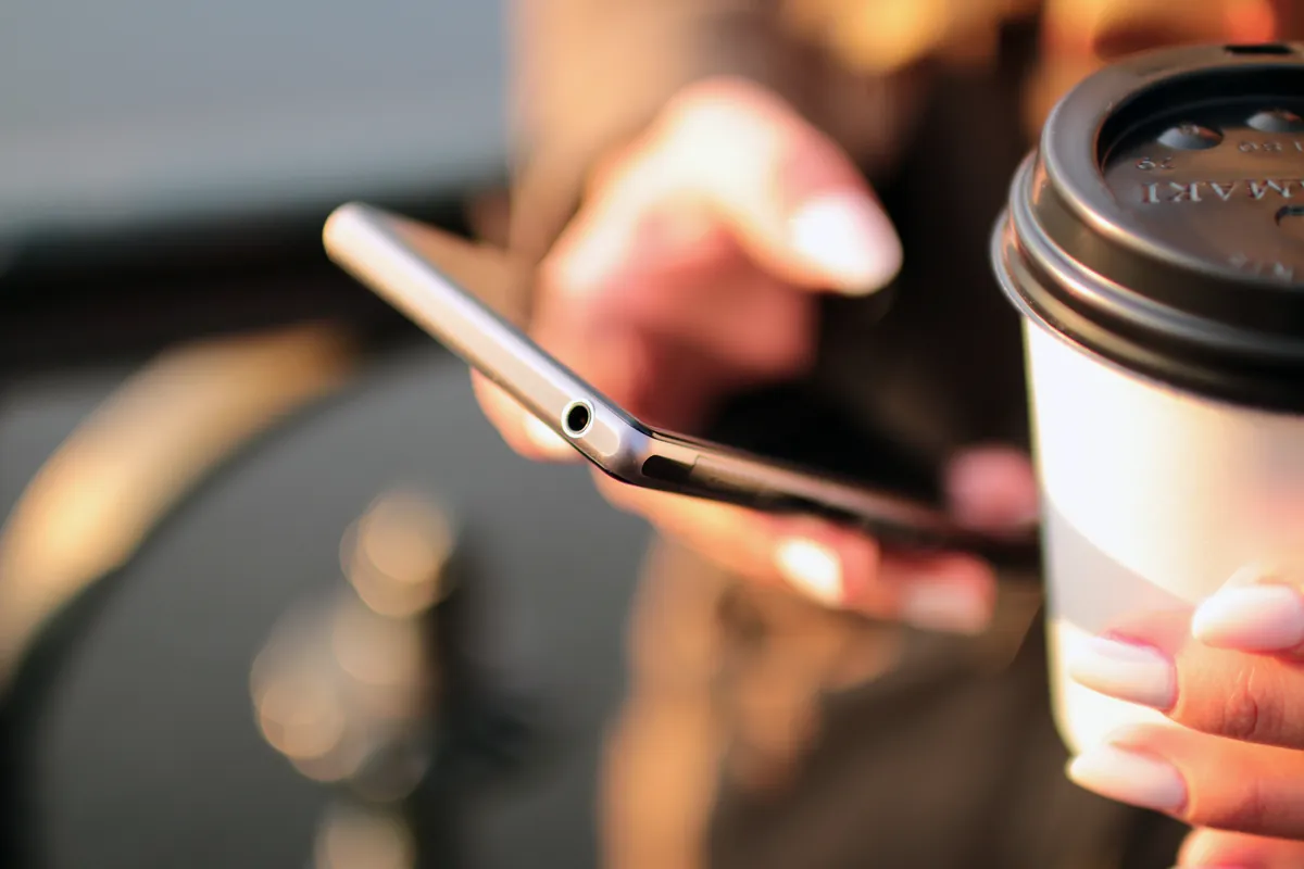 Hand hält ein Smartphone, daneben ein Kaffeebecher